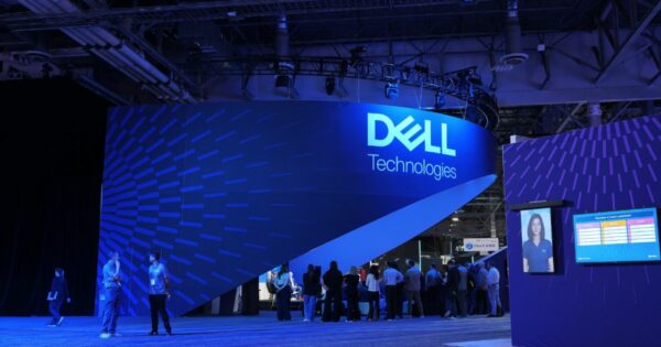 Grande stand da Dell Technologies numa feira tecnológica, iluminado em tons de azul. O logótipo da Dell Technologies está destacado numa parede curva e moderna, com várias pessoas a interagir e circular pelo espaço. À direita, vê-se um ecrã digital com informações e a imagem de uma colaboradora. (Nvidia, dados)