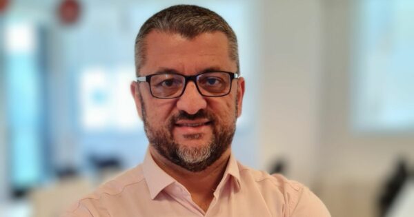 Homem de meia-idade com barba e cabelo curto, ambos grisalhos, usando óculos de armação preta e camisa social rosa clara. Ele está sorrindo levemente e olhando diretamente para a câmera. O fundo está desfocado, sugerindo um ambiente de escritório moderno e bem iluminado, com móveis e janelas visíveis de forma indistinta.