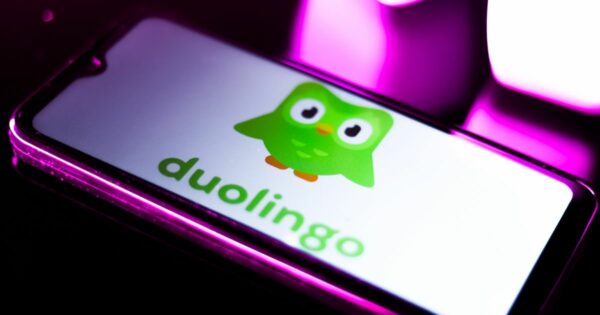Smartphone com o ecrã a exibir o logótipo do Duolingo, que consiste numa coruja verde estilizada com olhos grandes e expressão simpática, acompanhado da palavra 'duolingo' escrita em minúsculas com letras verdes. O telemóvel está iluminado por luzes de tonalidade rosa e roxa, criando um ambiente moderno e vibrante.