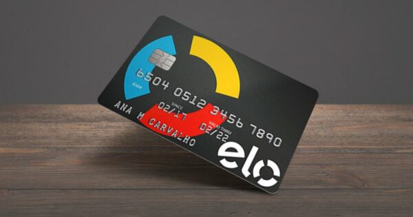 Cartão de crédito da bandeira Elo com fundo preto e elementos gráficos nas cores azul, amarelo e vermelho. O cartão está inclinado sobre uma superfície de madeira. Ele exibe um chip, o número do cartão, datas de validade (02/17 a 02/22) e o nome da titular, Ana M. Carvalho. O logotipo da Elo aparece em branco no canto inferior direito (solução)