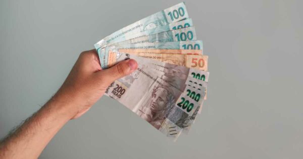 Mão segurando várias cédulas de real brasileiro em leque, com notas de 50, 100 e 200 reais visíveis, sobre fundo cinza neutro. (fintechs, open finance)