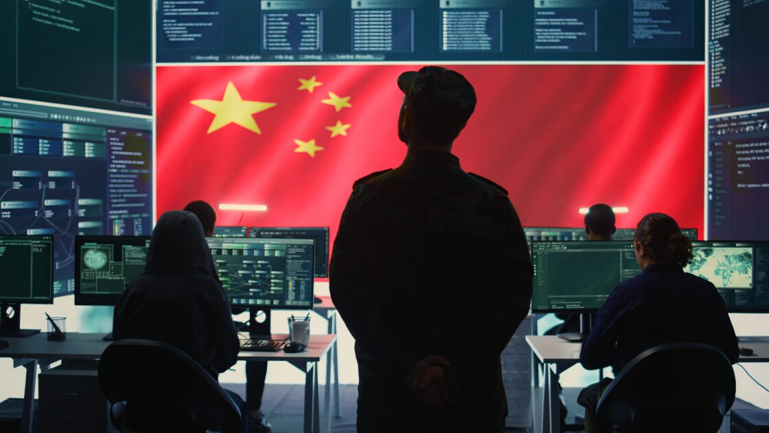 Uma pessoa está em frente a estações de computador e uma grande tela exibindo a bandeira chinesa, enquanto outros trabalham em mesas com vários monitores, apple, china