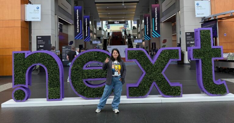 Andrea Ribeiro, Nutanix Technology Champion, posa sorridente em frente à escultura do evento ‘.NEXT’, feita com moldura roxa e preenchida por folhagens verdes. O cenário é o saguão de um centro de convenções, com banners do evento Nutanix .NEXT exibindo o slogan ‘Run Anything Anywhere’. Andrea veste uma camiseta amarela com a estampa ‘We can do it’, blazer listrado, calça jeans e crachá de participante.