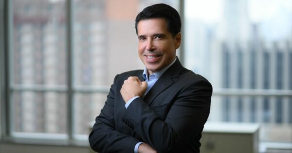 Carlos Perobelli, diretor-executivo da Nexmuv e idealizador da The Garage, posando sorridente, de terno escuro, em um ambiente corporativo com janelas amplas e vista desfocada de prédios ao fundo.