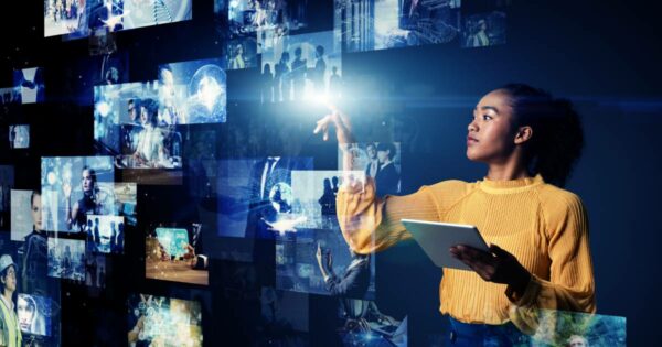 Imagem de uma mulher usando um tablet enquanto interage com uma interface virtual holográfica repleta de imagens e ícones flutuantes, representando conceitos de tecnologia, inteligência artificial e comunicação digital. A mulher veste uma blusa amarela de mangas longas e está em um ambiente de fundo escuro, com diversas telas translúcidas exibindo pessoas, gráficos e elementos tecnológicos (IA generativa, pensamento crítico, uso, inovação)
