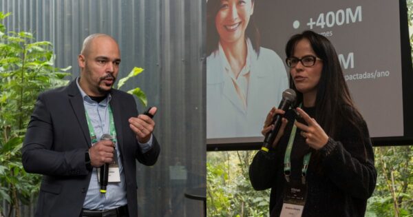 Marco Antonio, gerente de sistemas do Hcor, e Anaterra Oliveira, CIO da Dasa, falam em evento com microfone em mãos; ao fundo, há vegetação visível por uma parede de vidro e uma tela de apresentação com dados parciais sobre impacto anual. Ambos usam crachás do evento IT Forum Na Mata