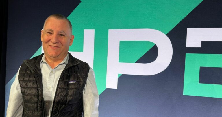 John Frey, diretor Sênior e Chief Technologist de Transformação Sustentável da HPE, está sorridente posando em frente a um painel com o logotipo da HPE estilizado em verde e branco sobre fundo escuro. Ele veste camisa social clara e colete preto da marca Patagonia.