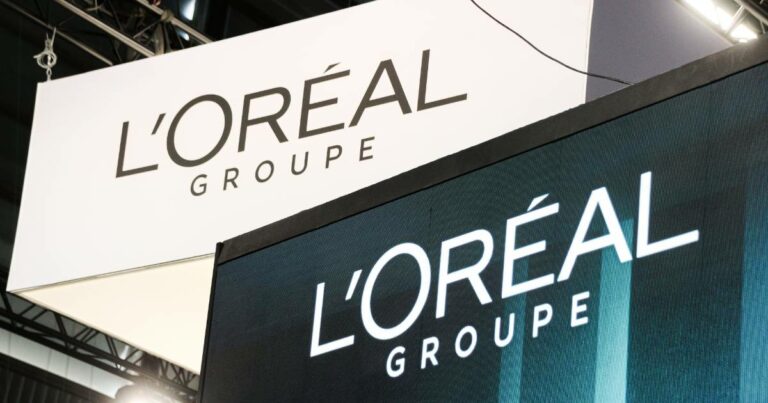 Imagem de dois painéis exibindo o logotipo da L'Oréal Groupe. O painel superior é branco com letras pretas, enquanto o painel inferior é uma tela digital com fundo escuro e letras brancas iluminadas. Ambos os painéis mostram o texto "L'ORÉAL GROUPE" em letras maiúsculas. A estrutura metálica do teto do local também é visível ao fundo