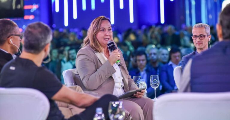 Marisa Reghini, vice-presidente de Negócios Digitais e Tecnologia do Banco do Brasil, participa de um painel de discussão em um evento. Ela está sentada em uma poltrona, segurando um microfone e um tablet, usando um terno bege com camisa branca. Ao seu redor, há outros participantes do painel e copos sobre a mesa. Ao fundo, um público atento e iluminação em tons de azul com luzes verticais destacam o ambiente moderno do evento.