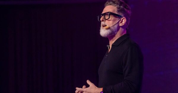 futuro: Neil Redding, CEO da Redding Futures e futurista, aparece em um palco durante uma palestra. Ele está de perfil, usando óculos de armação preta, microfone de cabeça e camisa preta de manga longa. O fundo é iluminado com luz roxa, criando um clima moderno e tecnológico.