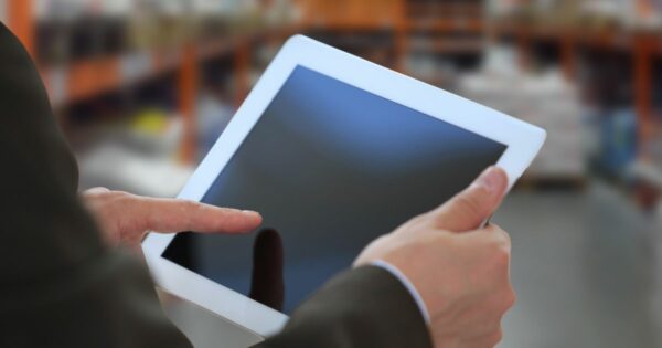 Uma pessoa segura um tablet branco com as duas mãos, enquanto toca a tela com o dedo indicador da mão direita. Ao fundo, o ambiente está desfocado, sugerindo um local interno como uma loja ou armazém, com prateleiras e diversos itens visíveis. (M.Dias Branco)