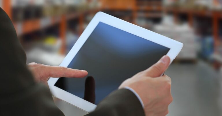 Uma pessoa segura um tablet branco com as duas mãos, enquanto toca a tela com o dedo indicador da mão direita. Ao fundo, o ambiente está desfocado, sugerindo um local interno como uma loja ou armazém, com prateleiras e diversos itens visíveis. (M.Dias Branco)
