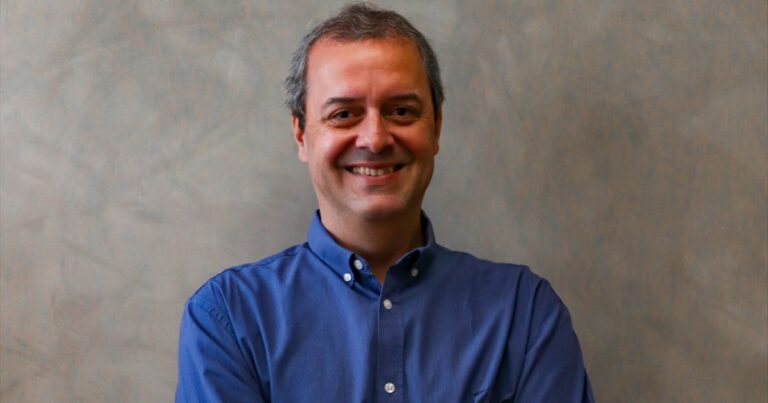 Homem de meia-idade com cabelos curtos e grisalhos, sorrindo com os braços cruzados. Ele veste uma camisa social azul e está posicionado diante de um fundo neutro em tom acinzentado, com textura suave. Sua expressão é simpática e acolhedora. (Serpro)