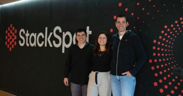 Três pessoasda Zup posam sorrindo em frente a uma parede preta com o logotipo da empresa 'StackSpot' em letras grandes brancas e elementos gráficos vermelhos em padrão de pontos. À esquerda está um homem de camisa preta, ao centro uma mulher com blusa preta e calça clara, e à direita um homem de moletom escuro e jeans claros. O ambiente parece ser corporativo e moderno.