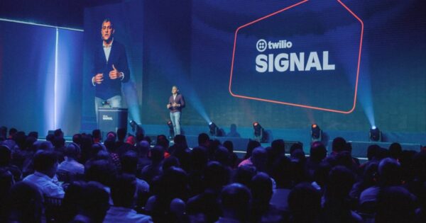 Um palestrante está no palco durante uma conferência ou evento, com o público sentado à frente. No fundo do palco, há uma grande tela exibindo o texto "twilio SIGNAL" junto com o logotipo da Twilio. À esquerda, uma tela menor mostra um close do palestrante.