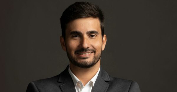 Homem jovem, de cabelos castanhos escuros e curtos, com barba bem aparada. Ele sorri levemente, veste um terno cinza escuro com camisa branca, e está posicionado contra um fundo liso, de tom escuro. Sua expressão transmite confiança e simpatia. (Intelbras)