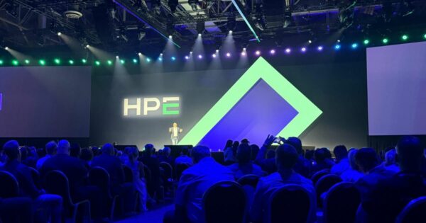 A imagem mostra Simon Ewington, vice‑presidente sênior de Canais e Ecossistema Global da HPE, em destaque no palco do HPE Discover 2025, em Las Vegas. Ele está de frente para um auditório lotado, gesticulando com as mãos, trajando blazer cinza claro e jeans. Atrás dele, um grande logo da HPE ocupa o centro da tela, iluminado por luzes azuis e verdes do evento, criando atmosfera vibrante e tecnológica. Sources