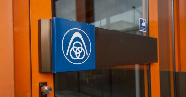 Detalhe de uma porta de vidro com estrutura metálica laranja, exibindo uma maçaneta horizontal com acabamento preto e um suporte lateral em metal escovado. No centro da maçaneta há uma placa azul com o logotipo da empresa Thyssenkrupp, que consiste em um arco estilizado acima de três elos entrelaçados, todos na cor branca. Ao fundo, é possível ver parte de um edifício e o reflexo da área externa.