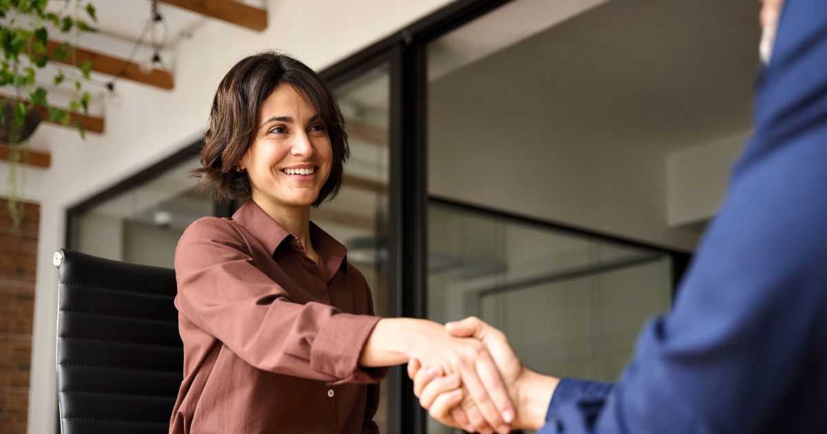 Mulher sorridente com cabelo castanho escuro e curto, vestindo uma camisa marrom, está sentada em uma cadeira de escritório e cumprimentando alguém com um aperto de mão. A cena parece ocorrer em um ambiente corporativo moderno, com paredes de vidro e vigas de madeira no teto. A pessoa com quem ela aperta a mão veste um terno azul, mas está fora de foco e parcialmente fora do enquadramento. A atmosfera transmite cordialidade e profissionalismo, sugerindo uma entrevista de emprego ou reunião de negócios. (vagas, candidato)