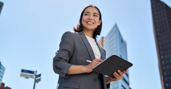 Descrição da imagem para o campo "Texto alternativo" do WordPress: Mulher executiva sorridente usando blazer cinza e blusa branca, segurando um tablet com uma caneta digital em uma mão. Ela está ao ar livre em um ambiente urbano, com prédios altos e modernos ao fundo, sob um céu azul claro e ensolarado. A perspectiva da foto é de baixo para cima, transmitindo sensação de confiança e profissionalismo (inovação)