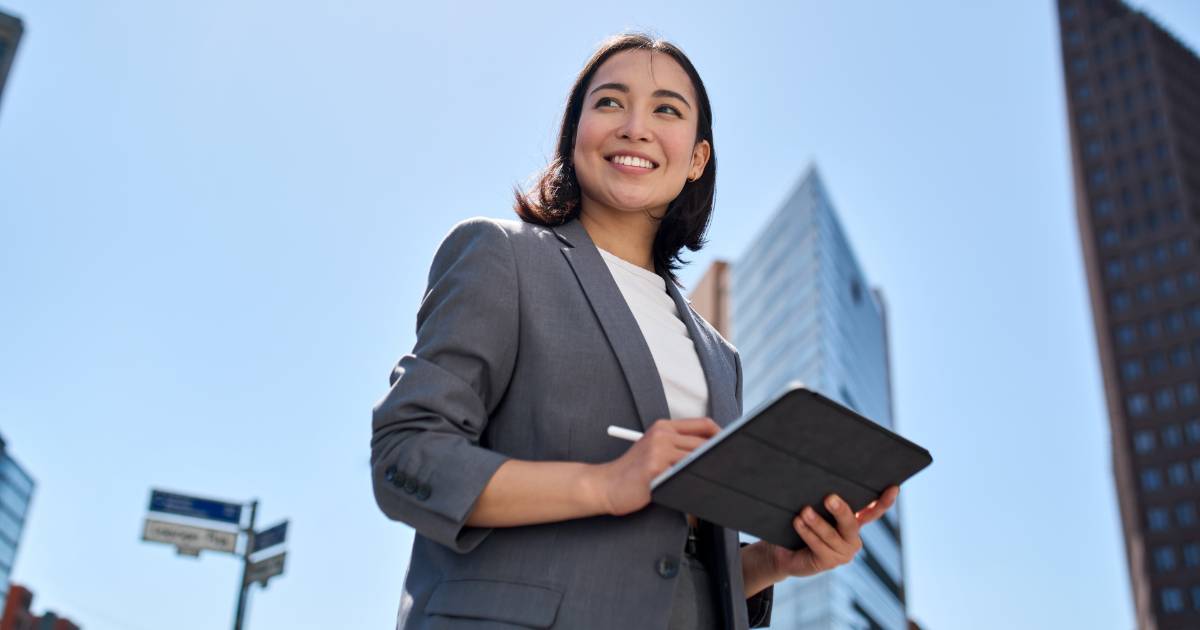 Descrição da imagem para o campo "Texto alternativo" do WordPress: Mulher executiva sorridente usando blazer cinza e blusa branca, segurando um tablet com uma caneta digital em uma mão. Ela está ao ar livre em um ambiente urbano, com prédios altos e modernos ao fundo, sob um céu azul claro e ensolarado. A perspectiva da foto é de baixo para cima, transmitindo sensação de confiança e profissionalismo (inovação)
