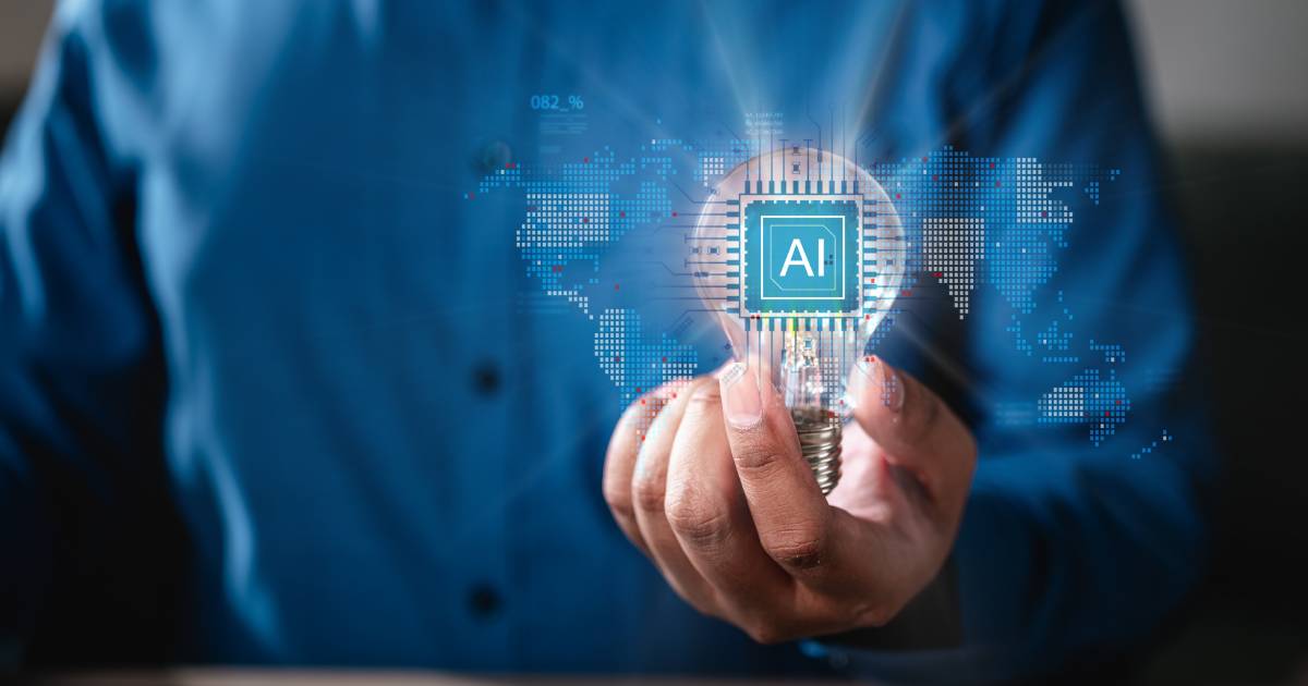 Pessoa vestindo camisa azul segura uma lâmpada acesa com o símbolo “AI” (inteligência artificial) no centro, estilizado como um chip de computador. Ao fundo, há uma representação digital do mapa-múndi com elementos gráficos que sugerem tecnologia e conectividade global, retorno, parceria (consciênte, startups, decisão, generativa, industria, processo, gratuitas, tendências, serviço)