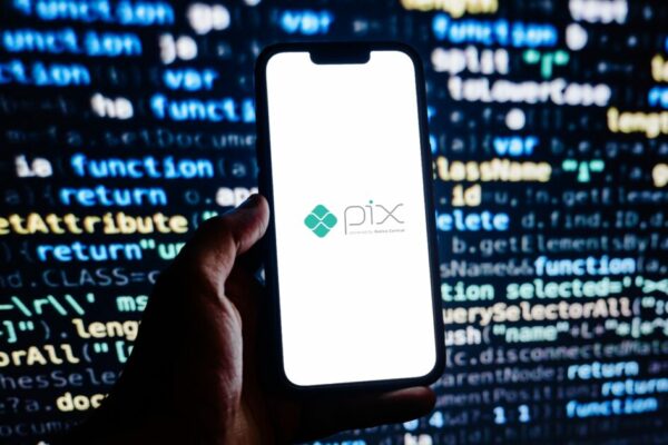 Pessoa segurando um smartphone exibindo a tela com o logo da plataforma PIX, enquanto o fundo mostra linhas de código em cores coloridas, destacando o tema de tecnologia e pagamentos digitais no Brasil
