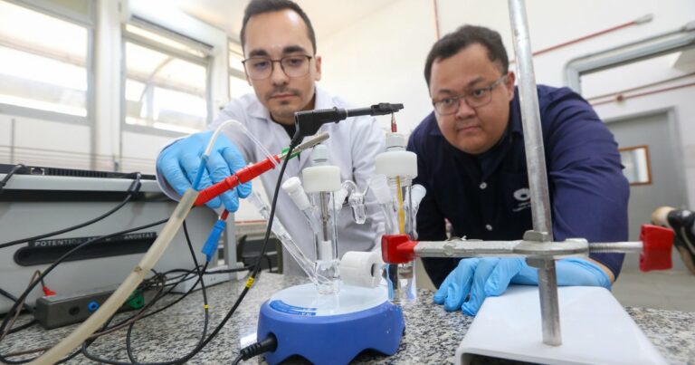 Imagem mostra dois pesquisadores em um laboratório da Unicamp manuseando equipamentos científicos. Ambos usam jalecos, luvas azuis e óculos de proteção. Um dos pesquisadores ajusta conexões elétricas em um aparato de vidro conectado a um agitador magnético azul sobre uma bancada. O ambiente é bem iluminado, com janelas amplas ao fundo e equipamentos laboratoriais visíveis, caracterizando uma atividade de pesquisa científica.