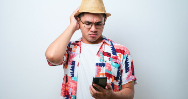 Imagem de um homem com expressão de confusão olhando para um celular. Ele está usando uma camisa estampada colorida, camiseta branca por baixo, chapéu de palha e óculos de grau. Sua mão esquerda está encostada na cabeça em um gesto de dúvida. O fundo é liso e branco (turista, viagem, viajante, férias)
