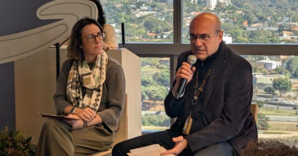 Imagem mostra Juliana Sztrajtman, presidente da Amazon.com.br, sentada à esquerda com um caderno no colo, usando óculos e um lenço estampado, e Cleber Morais, diretor-geral da AWS no Brasil, à direita, falando ao microfone e segurando folhas de papel. Ambos participam de um evento em ambiente interno com vista para uma área urbana ao fundo.