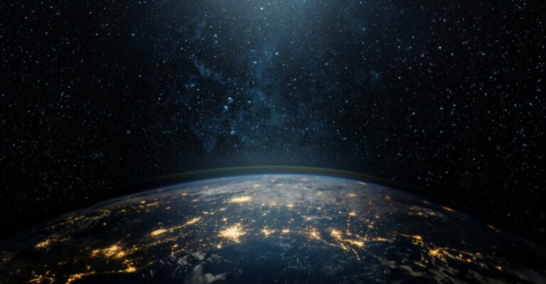 Imagem da Terra vista do espaço durante a noite, com a curvatura do planeta visível e diversas áreas iluminadas por luzes artificiais, representando grandes centros urbanos. Ao fundo, o céu escuro está repleto de estrelas, simbolizando a imensidão do universo e o conceito de globalização e conectividade mundial.