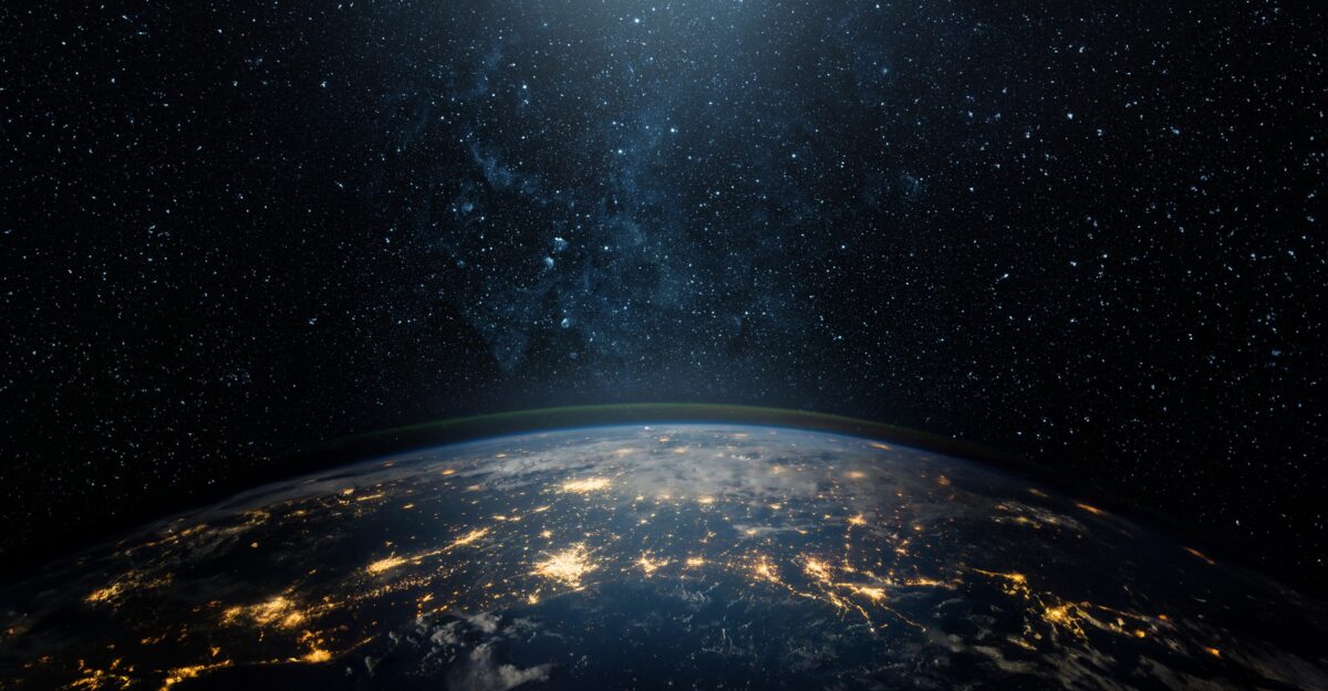 Imagem da Terra vista do espaço durante a noite, com a curvatura do planeta visível e diversas áreas iluminadas por luzes artificiais, representando grandes centros urbanos. Ao fundo, o céu escuro está repleto de estrelas, simbolizando a imensidão do universo e o conceito de globalização e conectividade mundial.