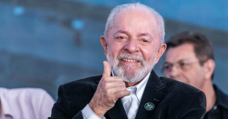 Homem idoso de barba branca e cabelos curtos e grisalhos, usando paletó preto com camisa branca, sorri e faz sinal de positivo com o polegar. Ele está em um ambiente público com outras pessoas ao fundo, levemente desfocadas. O homem veste um broche com o logo dos 30 anos da UNIFESP, indicando participação em evento relacionado à universidade (Lula, Luiz Inácio Lula da Silva, Brasil, Americanas, big techs, governo)