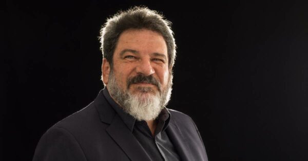 Retrato do filósofo e professor Mario Sergio Cortella, com fundo preto. Ele está com expressão séria e levemente sorridente, veste paletó escuro e camisa preta. Seu cabelo é grisalho e a barba cheia, também grisalha. A iluminação destaca seu rosto e parte superior do corpo, criando um efeito de contraste com o fundo escuro.