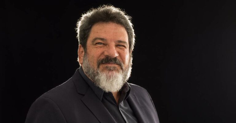 Retrato do filósofo e professor Mario Sergio Cortella, com fundo preto. Ele está com expressão séria e levemente sorridente, veste paletó escuro e camisa preta. Seu cabelo é grisalho e a barba cheia, também grisalha. A iluminação destaca seu rosto e parte superior do corpo, criando um efeito de contraste com o fundo escuro.