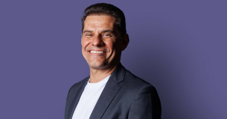 Homem sorridente de cabelos curtos e escuros, vestindo camiseta branca e blazer azul-marinho, posando sobre fundo roxo. Na imagem, está Paulo Secco, CEO da Mignow.