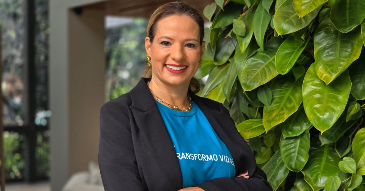 Uma pessoa está em pé com os braços cruzados, vestindo uma camisa azul com o texto parcialmente visível "TRANSFORMO VIDAS" e um blazer preto. O rosto está coberto por um bloco retangular. Ao fundo, há folhas verdes do lado direito e um ambiente interno desfocado do lado esquerdo. (profissionais, tecnologia)