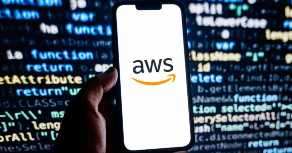 Uma mão segura um smartphone cuja tela exibe o logotipo da "AWS" (Amazon Web Services). Ao fundo, há linhas de código coloridas, sugerindo um ambiente de programação ou desenvolvimento de software.