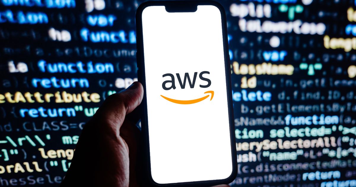 Uma mão segura um smartphone cuja tela exibe o logotipo da "AWS" (Amazon Web Services). Ao fundo, há linhas de código coloridas, sugerindo um ambiente de programação ou desenvolvimento de software.