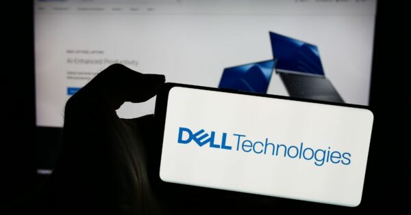 Uma mão segura um smartphone cuja tela exibe o logotipo da Dell Technologies. Ao fundo, desfocada, há uma tela de computador mostrando uma página da web com imagens de laptops azuis, sugerindo conteúdo relacionado à marca Dell.