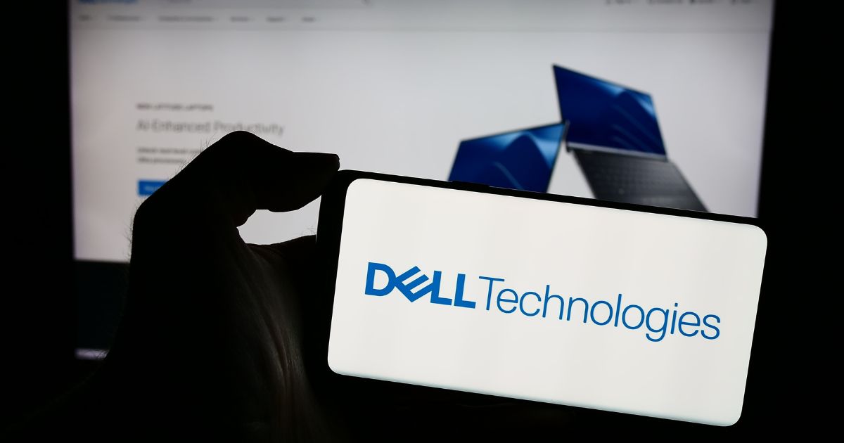 Uma mão segura um smartphone cuja tela exibe o logotipo da Dell Technologies. Ao fundo, desfocada, há uma tela de computador mostrando uma página da web com imagens de laptops azuis, sugerindo conteúdo relacionado à marca Dell.