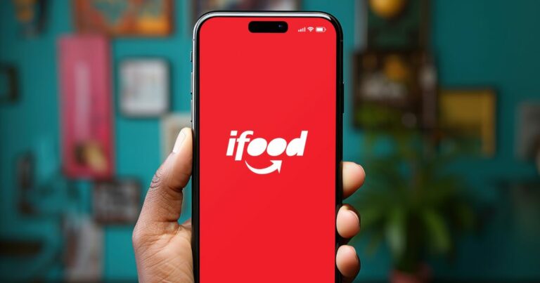 Uma mão segura um smartphone com o aplicativo iFood aberto na tela. O visor exibe o logotipo do iFood, composto pela palavra "iFood" em texto branco sobre um fundo vermelho, com uma seta estilizada em forma de sorriso abaixo. O fundo da imagem está desfocado, mas é possível identificar um ambiente interno com quadros emoldurados e uma planta.