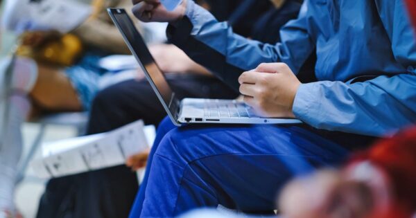 Uma pessoa está sentada utilizando um laptop. Ela veste calça e jaqueta azuis. Suas mãos estão posicionadas sobre o teclado, digitando. Ao fundo, há outras pessoas visíveis, algumas segurando papéis ou documentos, mas estão fora de foco. A atenção principal da imagem está nas mãos da pessoa e no laptop. (vagas e cursos e carreira)