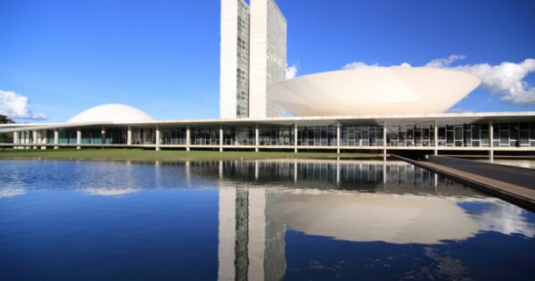 A imagem mostra o Congresso Nacional do Brasil, em Brasília. O complexo arquitetônico é composto por duas torres altas ladeadas por duas cúpulas — uma em cada lado. A estrutura está refletida em um espelho d’água no primeiro plano da imagem. (governo) para falar da estratégia nacional de transformação digital.