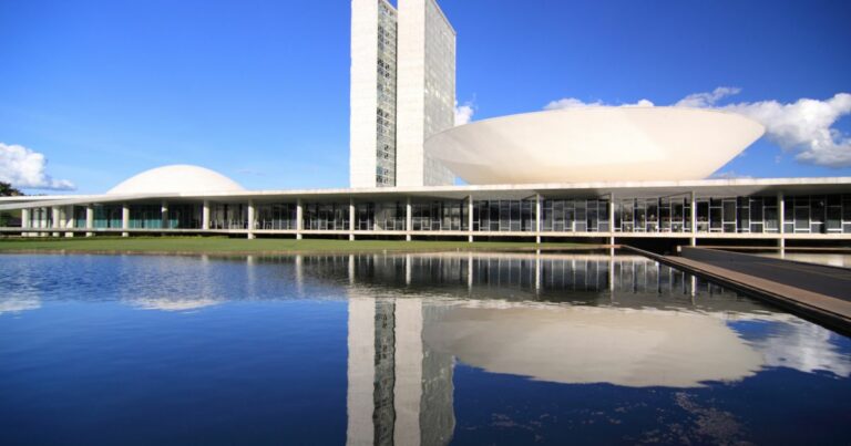 A imagem mostra o Congresso Nacional do Brasil, em Brasília. O complexo arquitetônico é composto por duas torres altas ladeadas por duas cúpulas — uma em cada lado. A estrutura está refletida em um espelho d’água no primeiro plano da imagem. (governo) para falar da estratégia nacional de transformação digital.