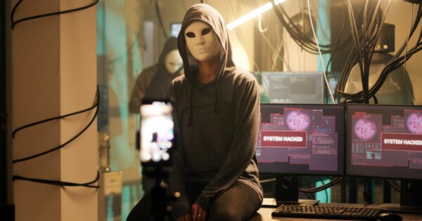 Uma pessoa com capuz está sentada em uma mesa em um ambiente com pouca iluminação, cercada por vários monitores de computador. O rosto da pessoa está pixelado, ocultando sua identidade. As telas exibem a mensagem "SYSTEM HACKED", acompanhada de linhas de código e gráficos. Há muitos cabos espalhados pelo local, reforçando a atmosfera de atividades cibernéticas. Uma câmera montada em um tripé está posicionada em frente à pessoa, sugerindo que ela pode estar gravando ou transmitindo algo. (Record)