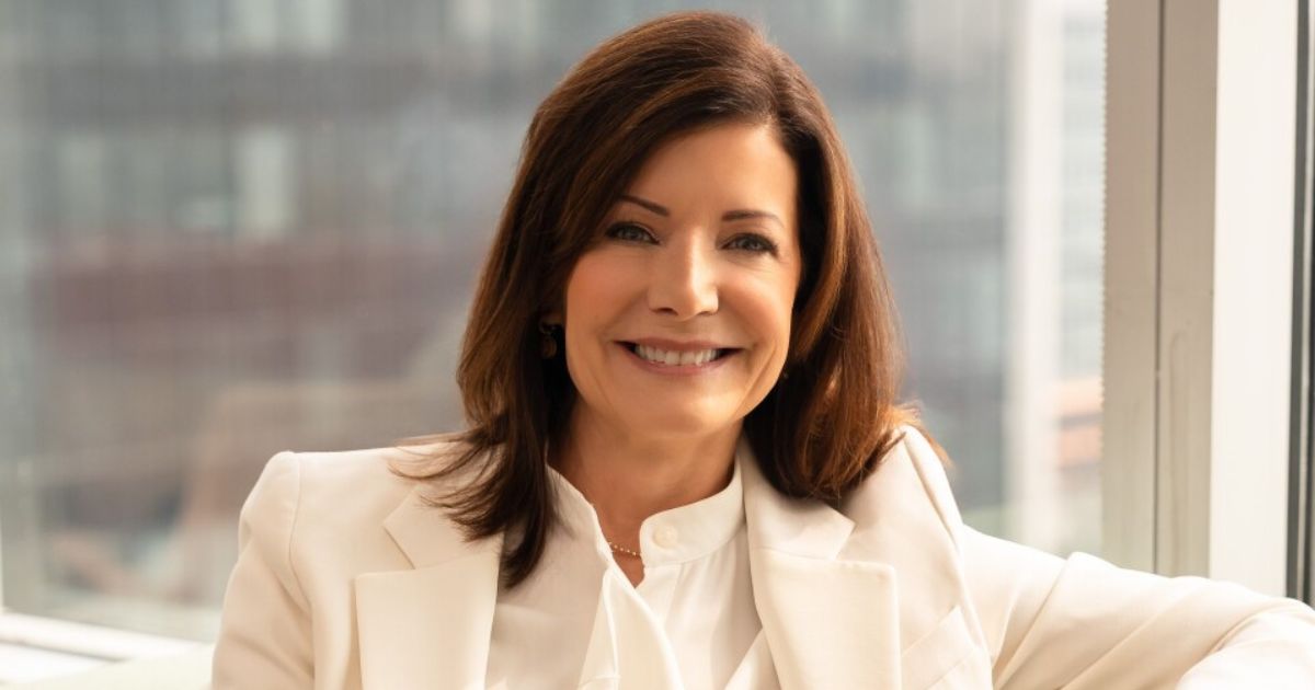 Arquelle Shaw, nova presidente Américas da Equinix, está sentada em um ambiente interno, vestindo um terno branco. Ela está em uma posição relaxada, com o braço direito apoiado em uma superfície. Ao fundo, há uma grande janela que mostra um cenário urbano desfocado.