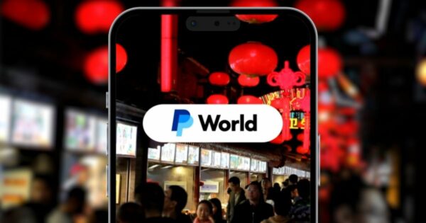 A imagem mostra a tela de um smartphone exibindo o logotipo do PayPal e a palavra "World" em destaque. Ao fundo, vê-se uma rua movimentada com pessoas e lanternas vermelhas penduradas, sugerindo um cenário típico de mercado ou festival asiático.