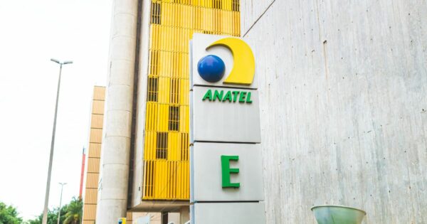 A imagem mostra uma placa em frente a um edifício. A placa exibe o logotipo e o nome "ANATEL", com uma esfera azul e uma forma de crescente amarela acima. Abaixo de "ANATEL", há uma grande letra "E" na cor verde. Ao fundo, o prédio possui painéis verticais amarelos e paredes de concreto.