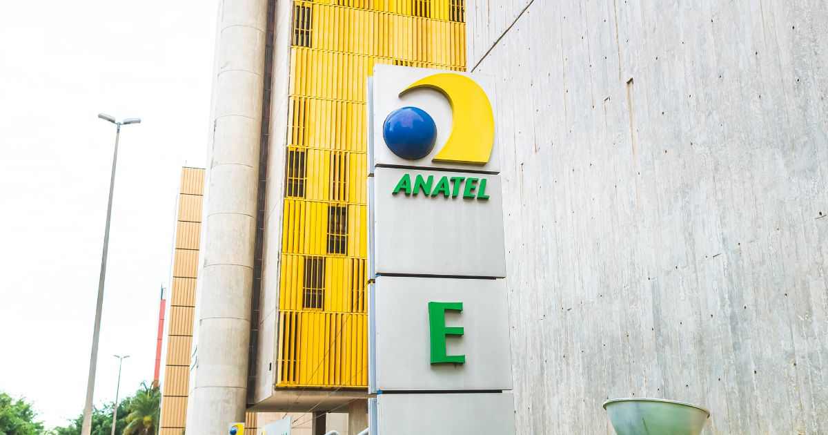A imagem mostra uma placa em frente a um edifício. A placa exibe o logotipo e o nome "ANATEL", com uma esfera azul e uma forma de crescente amarela acima. Abaixo de "ANATEL", há uma grande letra "E" na cor verde. Ao fundo, o prédio possui painéis verticais amarelos e paredes de concreto.